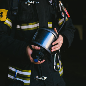 Firefighter standing with protective gear and gas mask, wearing a uniform in a dark setting.